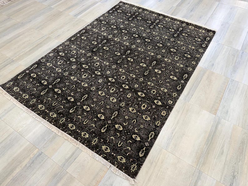 Puede incluir: Una alfombra rectangular con un fondo gris carb&oacute;n oscuro y un patr&oacute;n repetitivo de motivos florales geom&eacute;tricos en color crema y beige claro. La alfombra tiene una textura de pelo corto y est&aacute; rematada con flecos claros en ambos extremos.