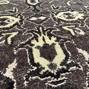Puede incluir: Una alfombra gruesa y texturizada con un fondo marr&oacute;n oscuro y un patr&oacute;n repetitivo de motivos florales y geom&eacute;tricos estilizados en color crema. La construcci&oacute;n de pelo largo crea una superficie suave y densa con detalles en contraste.