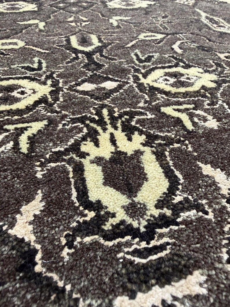 Puede incluir: Una alfombra gruesa y texturizada con un fondo marr&oacute;n oscuro y un patr&oacute;n repetitivo de motivos florales y geom&eacute;tricos estilizados en color crema. La construcci&oacute;n de pelo largo crea una superficie suave y densa con detalles en contraste.