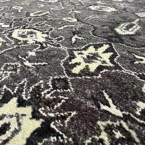 Puede incluir: Una alfombra gruesa y texturizada con un fondo gris carb&oacute;n oscuro y un intrincado patr&oacute;n floral y geom&eacute;trico repetitivo en crema y negro. El pelo es denso y afelpado, mostrando un dise&ntilde;o de alto contraste con detalles de mechones visibles.