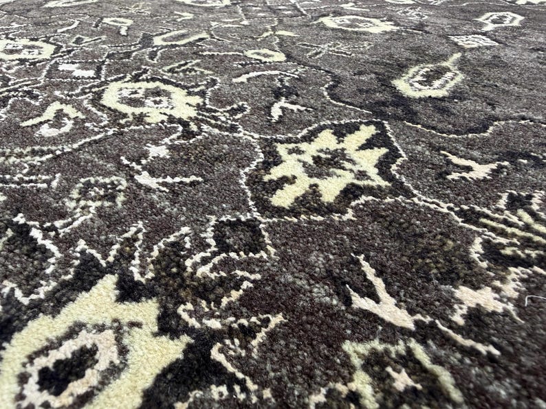 Puede incluir: Una alfombra gruesa y texturizada con un fondo gris carb&oacute;n oscuro y un intrincado patr&oacute;n floral y geom&eacute;trico repetitivo en crema y negro. El pelo es denso y afelpado, mostrando un dise&ntilde;o de alto contraste con detalles de mechones visibles.