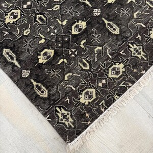 Puede incluir: Una alfombra de color gris carb&oacute;n oscuro con un patr&oacute;n geom&eacute;trico repetitivo en crema y beige claro. La alfombra tiene un pelo denso y texturizado, y est&aacute; rematada con flecos cortos de color crema a lo largo del borde recto del tejido.