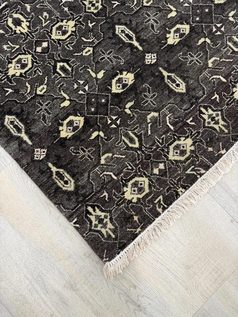 Puede incluir: Una alfombra de color gris carb&oacute;n oscuro con un patr&oacute;n geom&eacute;trico repetitivo en crema y beige claro. La alfombra tiene un pelo denso y texturizado, y est&aacute; rematada con flecos cortos de color crema a lo largo del borde recto del tejido.