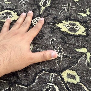 Puede incluir: Una alfombra de color gris carb&oacute;n oscuro con un patr&oacute;n repetitivo de motivos florales y geom&eacute;tricos en color crema claro. La superficie de la alfombra tiene un pelo denso y texturizado con fibras visibles en los dise&ntilde;os.