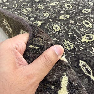 Puede incluir: Una alfombra de color marr&oacute;n oscuro con un patr&oacute;n geom&eacute;trico repetitivo en color crema claro. La alfombra tiene una textura de pelo grueso y tupido, y el borde est&aacute; doblado para revelar la construcci&oacute;n densa y el reverso del material textil.