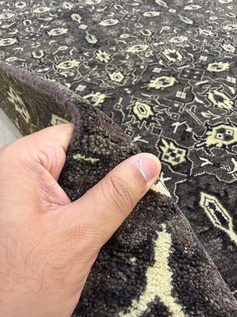 Puede incluir: Una alfombra de color marr&oacute;n oscuro con un patr&oacute;n geom&eacute;trico repetitivo en color crema claro. La alfombra tiene una textura de pelo grueso y tupido, y el borde est&aacute; doblado para revelar la construcci&oacute;n densa y el reverso del material textil.