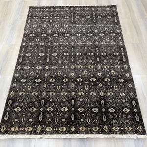 Puede incluir: Una alfombra rectangular con un fondo gris oscuro y negro que presenta un intrincado patr&oacute;n geom&eacute;trico y floral repetitivo en color crema. La alfombra tiene un pelo corto y denso y est&aacute; rematada con flecos blancos en los bordes.