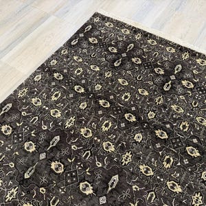 Puede incluir: Una alfombra rectangular con un fondo gris carb&oacute;n oscuro y un patr&oacute;n repetitivo de motivos geom&eacute;tricos y florales de color crema. La alfombra tiene una textura de pelo corto e incluye un fleco de color claro en el borde superior.
