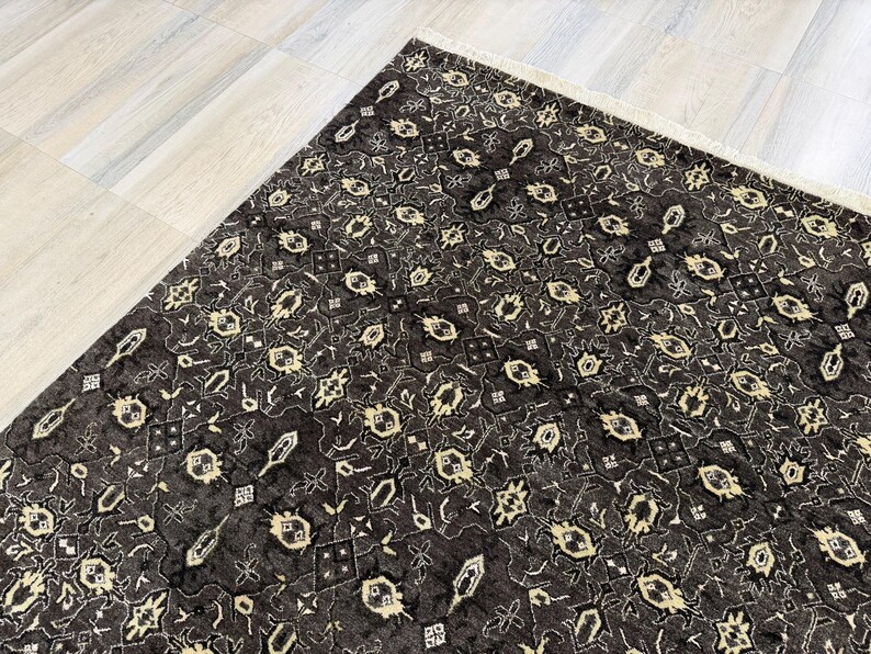 Puede incluir: Una alfombra rectangular con un fondo gris carb&oacute;n oscuro y un patr&oacute;n repetitivo de motivos geom&eacute;tricos y florales de color crema. La alfombra tiene una textura de pelo corto e incluye un fleco de color claro en el borde superior.