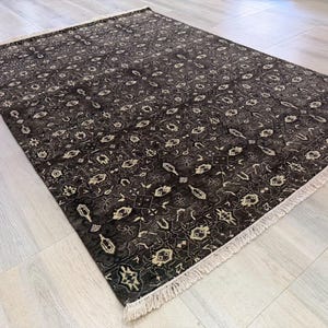 Puede incluir: Una alfombra rectangular con fondo marr&oacute;n oscuro y un intrincado patr&oacute;n geom&eacute;trico repetitivo en crema y beige claro. La alfombra tiene una textura de pelo corto y est&aacute; rematada con flecos de color crema en los dos extremos m&aacute;s cortos.