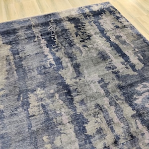 Puede incluir: Una alfombra rectangular con un patr&oacute;n abstracto y texturizado en tonos azul marino profundo, gris pizarra y plata claro. El dise&ntilde;o consiste en rayas verticales de aspecto desgastado y parches moteados que crean una apariencia en capas.