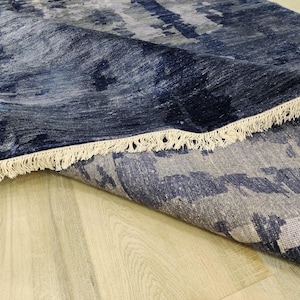 Puede incluir: Una alfombra rectangular con un patr&oacute;n abstracto en tonos azul marino profundo, gris pizarra y blanco. La alfombra tiene una esquina doblada que revela una superficie texturizada y est&aacute; rematada con flecos de color blanco roto.