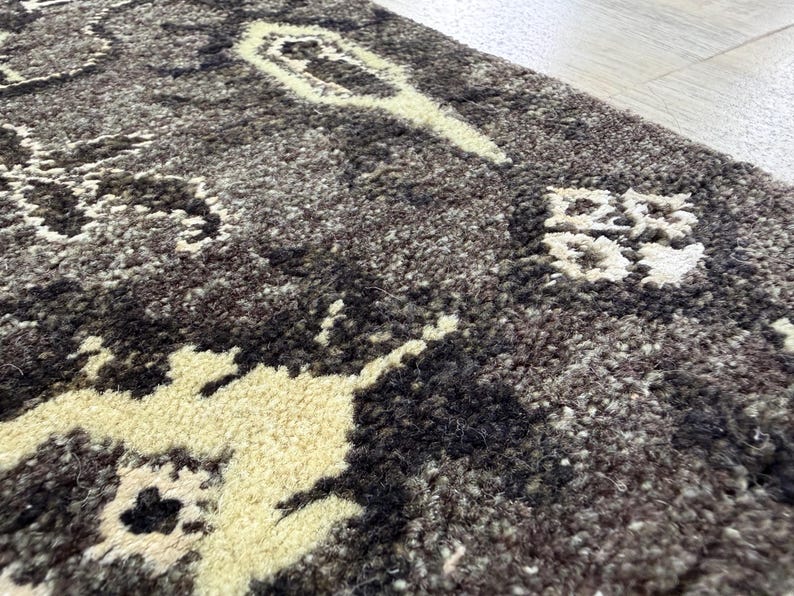 Puede incluir: Una alfombra gruesa y texturizada con una base de color gris carb&oacute;n oscuro y marr&oacute;n oscuro, con patrones abstractos florales y geom&eacute;tricos de color crema tejidos en el pelo denso y afelpado de la superficie.