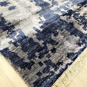 Puede incluir: Una alfombra rectangular con un patr&oacute;n abstracto en tonos azul marino, gris pizarra y blanco. La alfombra tiene una superficie de pelo texturizado y est&aacute; rematada con una hilera de flecos de color crema a lo largo del borde corto.