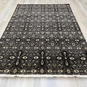 Puede incluir: Una alfombra rectangular con un fondo gris carb&oacute;n oscuro y un intrincado patr&oacute;n geom&eacute;trico y floral repetitivo en crema y gris claro. La alfombra presenta un fleco corto de color blanco a lo largo del borde inferior y textura baja.