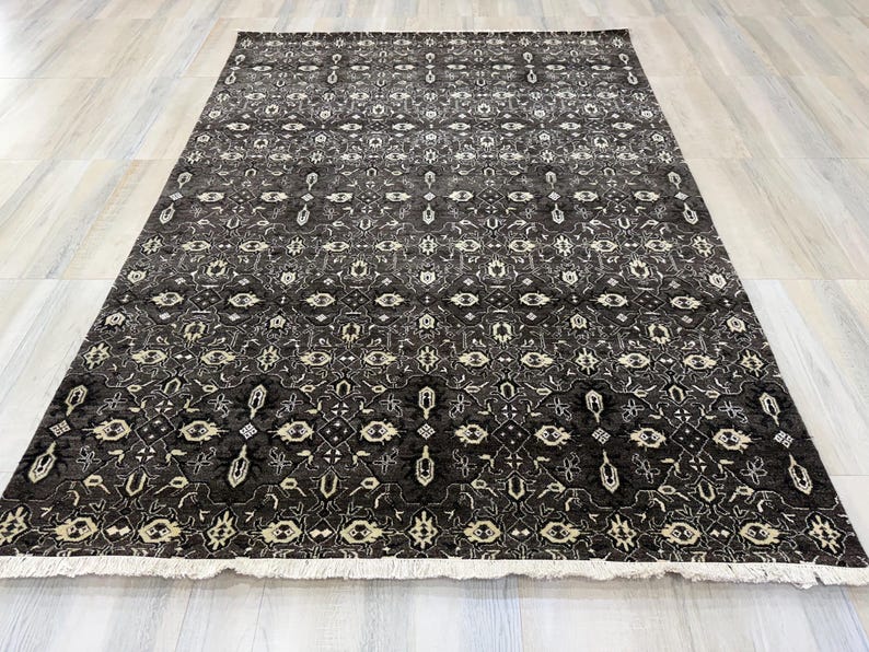 Puede incluir: Una alfombra rectangular con un fondo gris carb&oacute;n oscuro y un intrincado patr&oacute;n geom&eacute;trico y floral repetitivo en crema y gris claro. La alfombra presenta un fleco corto de color blanco a lo largo del borde inferior y textura baja.
