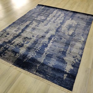 Puede incluir: Una alfombra rectangular con un patr&oacute;n abstracto en tonos azul marino, azul pizarra y gris claro. El dise&ntilde;o consiste en rayas verticales texturizadas y manchas de color. La alfombra tiene flecos cortos de color claro en ambos extremos.