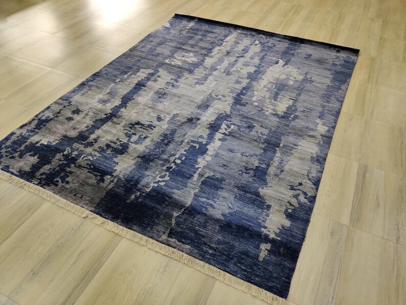 Puede incluir: Una alfombra rectangular con un patr&oacute;n abstracto en tonos azul marino, azul pizarra y gris claro. El dise&ntilde;o consiste en rayas verticales texturizadas y manchas de color. La alfombra tiene flecos cortos de color claro en ambos extremos.