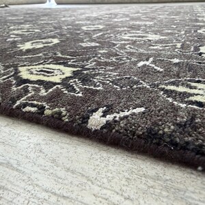 Puede incluir: Una alfombra de color marr&oacute;n oscuro con un patr&oacute;n intrincado y repetitivo en crema y negro. La alfombra tiene un pelo grueso y texturizado con un borde rematado, mostrando un motivo detallado inspirado en flores en toda la superficie.