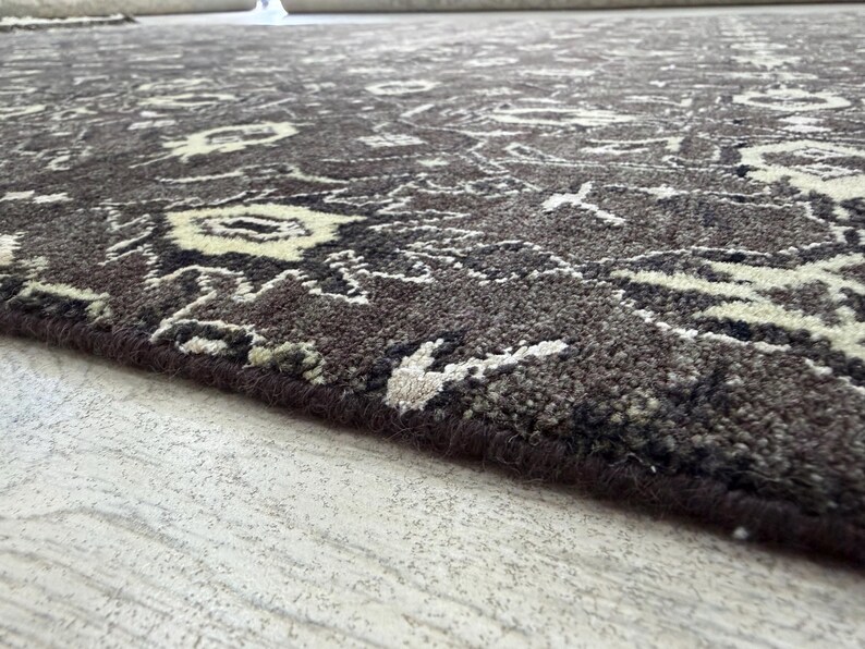Puede incluir: Una alfombra de color marr&oacute;n oscuro con un patr&oacute;n intrincado y repetitivo en crema y negro. La alfombra tiene un pelo grueso y texturizado con un borde rematado, mostrando un motivo detallado inspirado en flores en toda la superficie.