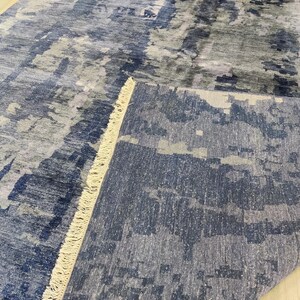 Puede incluir: Una alfombra rectangular con un patr&oacute;n abstracto en tonos azul marino, gris pizarra y blanco. La alfombra tiene una esquina doblada que revela una superficie texturizada y una hilera de flecos cortos de color crema a lo largo del pliegue.