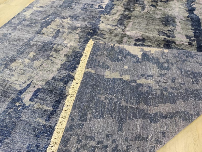 Puede incluir: Una alfombra rectangular con un patr&oacute;n abstracto en tonos azul marino, gris pizarra y blanco. La alfombra tiene una esquina doblada que revela una superficie texturizada y una hilera de flecos cortos de color crema a lo largo del pliegue.