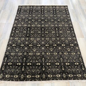 Puede incluir: Una alfombra rectangular con un fondo gris oscuro y negro que presenta un patr&oacute;n intrincado y repetitivo en color crema y gris claro. La alfombra tiene un dise&ntilde;o geom&eacute;trico y floral sim&eacute;trico en toda su superficie y flecos blancos cortos.