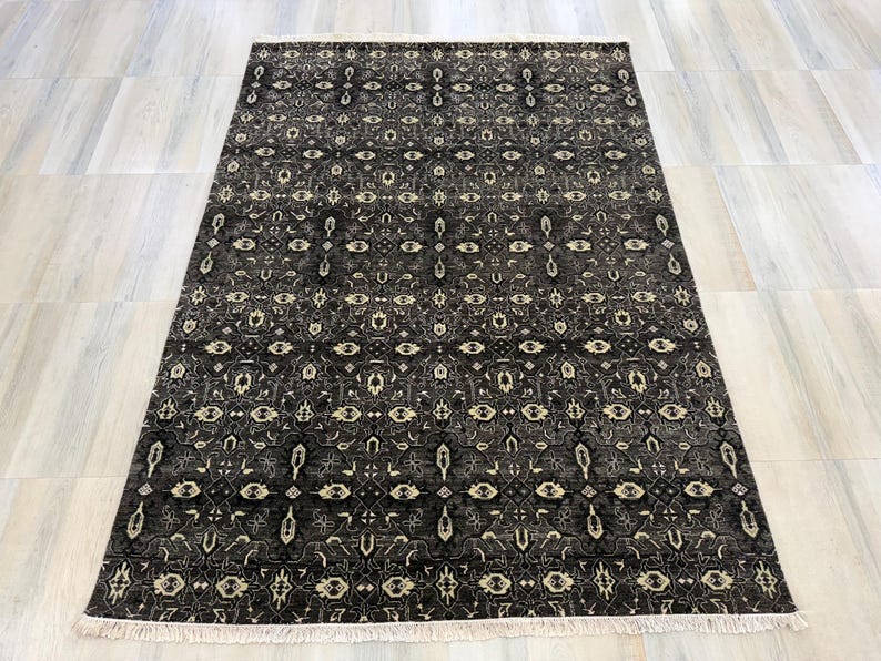 Puede incluir: Una alfombra rectangular con un fondo gris oscuro y negro que presenta un patr&oacute;n intrincado y repetitivo en color crema y gris claro. La alfombra tiene un dise&ntilde;o geom&eacute;trico y floral sim&eacute;trico en toda su superficie y flecos blancos cortos.
