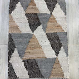 Puede incluir: Una alfombra rectangular con un patrón geométrico de triángulos en tonos grises, marrones y blancos. La alfombra tiene un borde con flecos y está hecha de material tejido. El diseño presenta un patrón repetitivo de triángulos.
