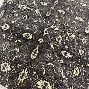 Puede incluir: Una alfombra de color gris carb&oacute;n oscuro con un patr&oacute;n geom&eacute;trico repetitivo en crema p&aacute;lido. La alfombra tiene un pelo denso y texturizado, con una esquina doblada que revela un fleco blanco en el borde. El dise&ntilde;o consta de motivos florales.