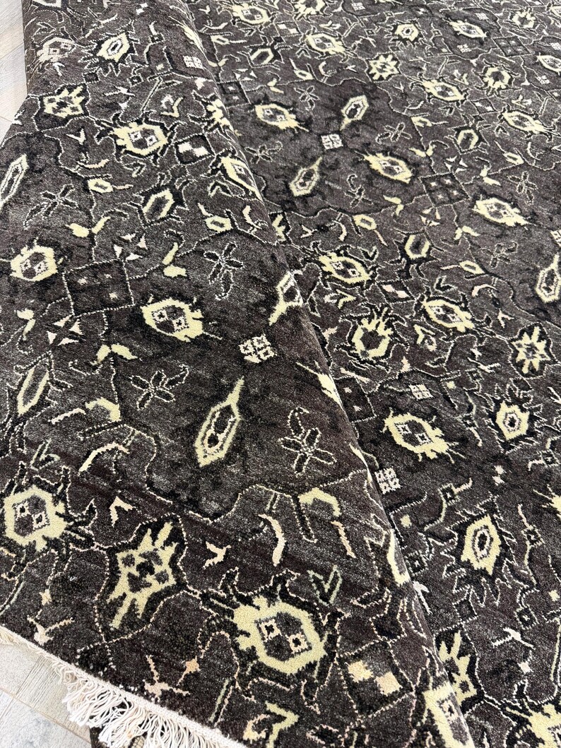 Puede incluir: Una alfombra de color gris carb&oacute;n oscuro con un patr&oacute;n geom&eacute;trico repetitivo en crema p&aacute;lido. La alfombra tiene un pelo denso y texturizado, con una esquina doblada que revela un fleco blanco en el borde. El dise&ntilde;o consta de motivos florales.