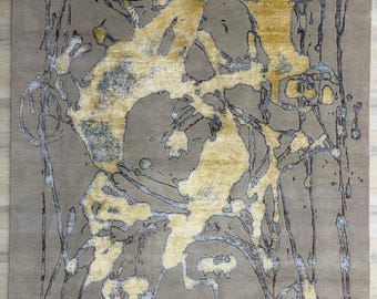 Puede incluir: Una alfombra rectangular con una base color topo y un patrón abstracto de salpicaduras y goteos de pintura en tonos dorados y gris oscuro. La alfombra tiene flecos cortos en los bordes superior e inferior, con una superficie texturizada.