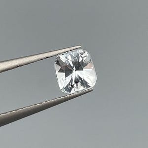 以下が含まれることがあります： 銀色のピンセットで保持された、透明な正方形カットの宝石。 宝石は光を反射するファセット加工された表面を持ち、きらめく効果を生み出しています。 背景は無地のニュートラルグレーです。