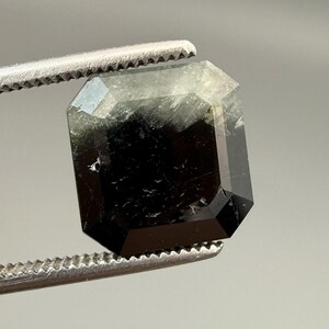 May include: A faceted, emerald-cut gemstone with a dark, smoky appearance. The gem is held by a pair of silver tweezers, showcasing its geometric shape and internal clarity. The stone's color transitions from black to a lighter, translucent hue.