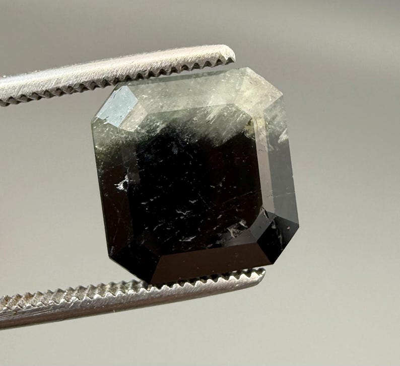 May include: A faceted, emerald-cut gemstone with a dark, smoky appearance. The gem is held by a pair of silver tweezers, showcasing its geometric shape and internal clarity. The stone's color transitions from black to a lighter, translucent hue.