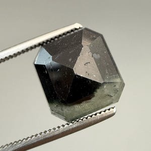 May include: A faceted, dark green gemstone held by a pair of silver tweezers. The gem has a geometric, hexagonal shape with multiple reflective surfaces. The stone appears translucent with a subtle, internal glow.