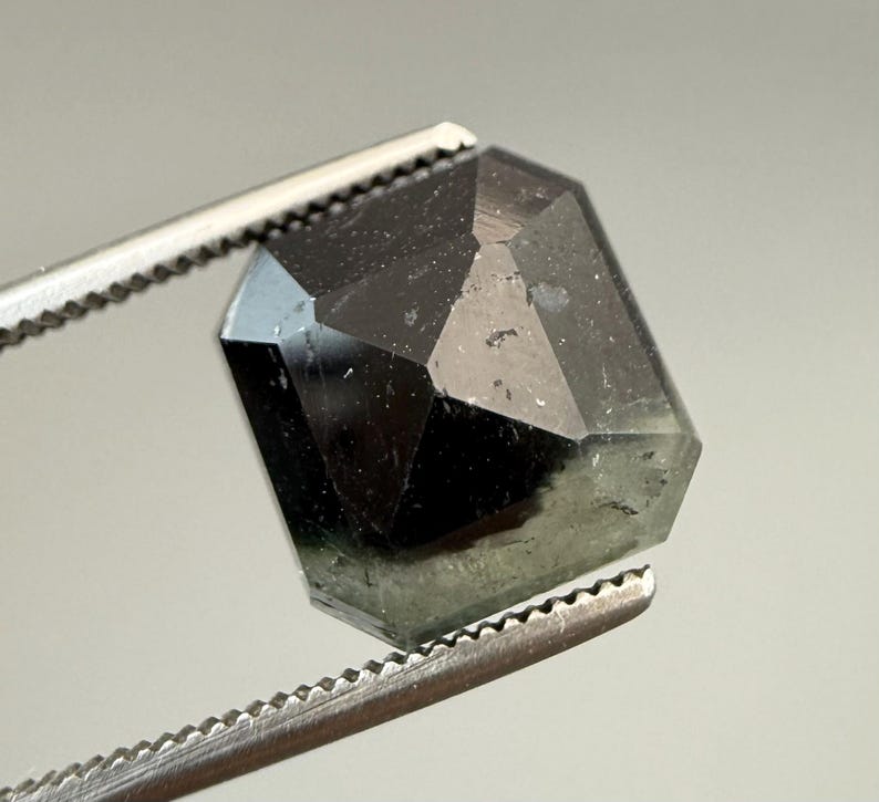 May include: A faceted, dark green gemstone held by a pair of silver tweezers. The gem has a geometric, hexagonal shape with multiple reflective surfaces. The stone appears translucent with a subtle, internal glow.