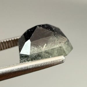 May include: A faceted, dark gemstone with a mix of black and green hues is held by a pair of tweezers. The gem's cut reveals multiple facets, creating a geometric shape. The tweezers are made of a shiny, silver metal.