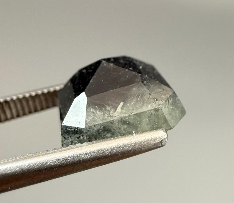 May include: A faceted, dark gemstone with a mix of black and green hues is held by a pair of tweezers. The gem's cut reveals multiple facets, creating a geometric shape. The tweezers are made of a shiny, silver metal.