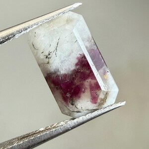 May include: A faceted, clear quartz crystal with a vibrant, deep red and pink tourmaline inclusion. The crystal is held by silver tweezers, showcasing its natural beauty and unique color variations. The gemstone is a striking example of natural mineral formations.