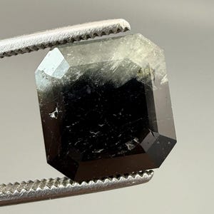 May include: A faceted, emerald-cut gemstone held by a pair of tweezers. The gem is primarily black with areas of translucent green and white. The facets reflect light, creating a sparkling effect. The tweezers are silver.