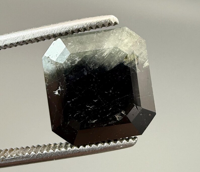 May include: A faceted, emerald-cut gemstone held by a pair of tweezers. The gem is primarily black with areas of translucent green and white. The facets reflect light, creating a sparkling effect. The tweezers are silver.