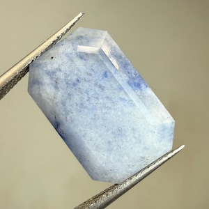 Peut inclure: Une pierre précieuse rectangulaire, facettée, de couleur bleu pâle et blanche. Elle est maintenue par une pince métallique, révélant ses surfaces polies et ses inclusions internes. La forme de la pierre est un prisme long à six faces.