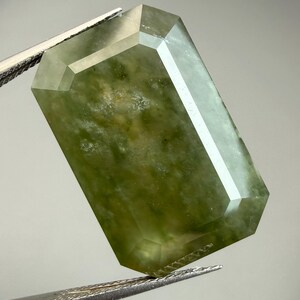 Peut inclure: Une pierre précieuse rectangulaire à facettes de couleur vert pâle. La gemme est maintenue par une pince, mettant en valeur ses surfaces polies et ses motifs internes. La forme de la pierre est un rectangle aux coins coupés.