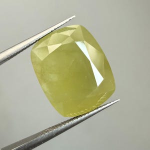 Könnte beinhalten: Ein facettierter, hellgelber Edelstein, der von einer Pinzette gehalten wird. Der Stein hat einen rechteckigen Kissenschliff mit mehreren Facetten, die das Licht reflektieren. Die Farbe ist ein blasses, fast chartreusefarbenes Gelb.