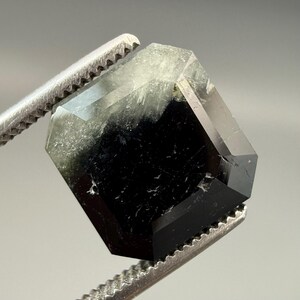 May include: A faceted, hexagonal gemstone held by a metal tweezer. The gem transitions from a clear, light green hue at the top to a deep black at the bottom. The facets reflect light, showcasing the gem's crystalline structure.