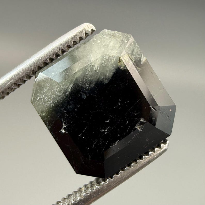 May include: A faceted, hexagonal gemstone held by a metal tweezer. The gem transitions from a clear, light green hue at the top to a deep black at the bottom. The facets reflect light, showcasing the gem's crystalline structure.