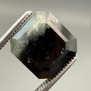 May include: A faceted, dark green and black gemstone held by a pair of silver tweezers. The gem has a geometric shape with multiple facets, reflecting light. The stone appears translucent in some areas.