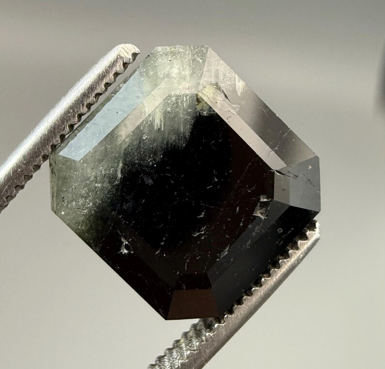 May include: A faceted, dark green and black gemstone held by a pair of silver tweezers. The gem has a geometric shape with multiple facets, reflecting light. The stone appears translucent in some areas.