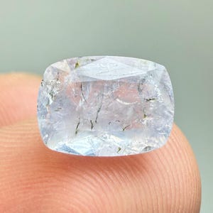 Peut inclure: Une pierre précieuse taille coussin, de couleur lavande pâle, présentant une surface à facettes et des inclusions sombres en forme d'aiguilles visibles dans tout son corps translucide. La pierre affiche une teinte violet clair avec des fractures internes.